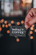 Hand holding a 'Impact Coffee' card with blurred lights in the background