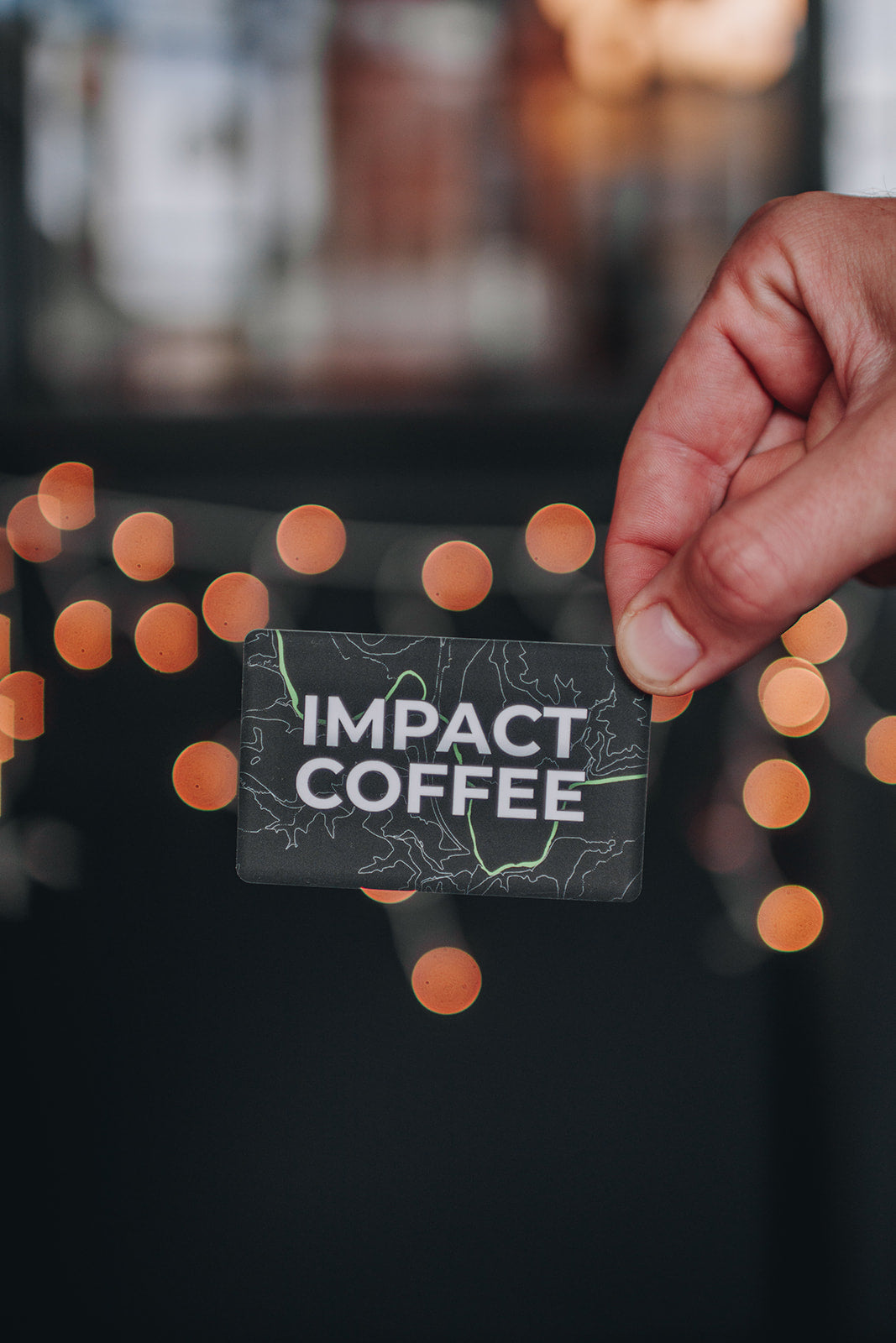 Hand holding a 'Impact Coffee' card with blurred lights in the background