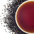 A cup of Irish Breakfast black tea with a teaspoon, surrounded by loose tea leaves and bits of dried tea on a white background.